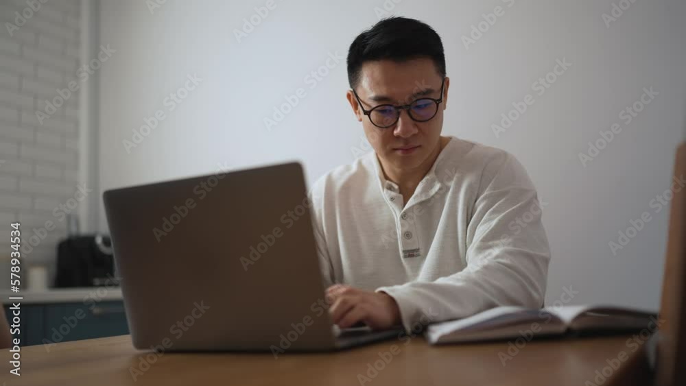 Serious brunet Asian man wearing eyeglasses working on laptop in the kitchen at home Serious brunet Asian man wearing eyeglasses working on laptop in the kitchen at home