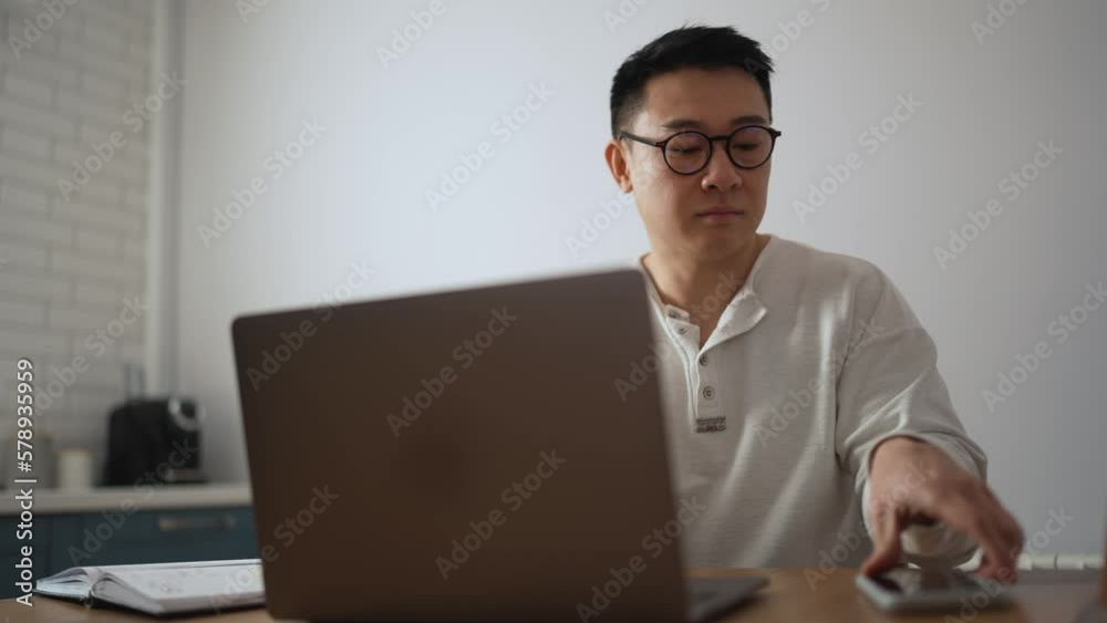 Pensive brunet Asian man wearing eyeglasses working on laptop and talking on mobile in the kitchen at home Pensive brunet Asian man wearing eyeglasses working on laptop and talking on mobile in the kitchen at home