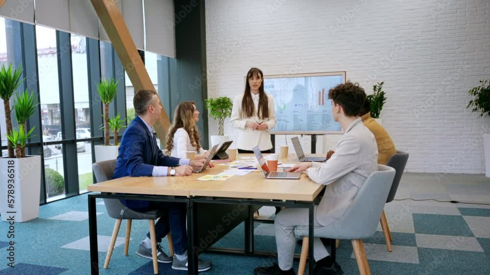 Female team leader at business meeting in an office, discussing business affairs with workers using a big display with charts Female team leader at business meeting in an office, discussing business affairs with workers using a big display with charts