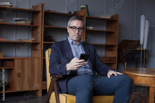 Hansome elderly businessman in blue suit and glasses working and looking on his mobile phone on office wall with boockshelf