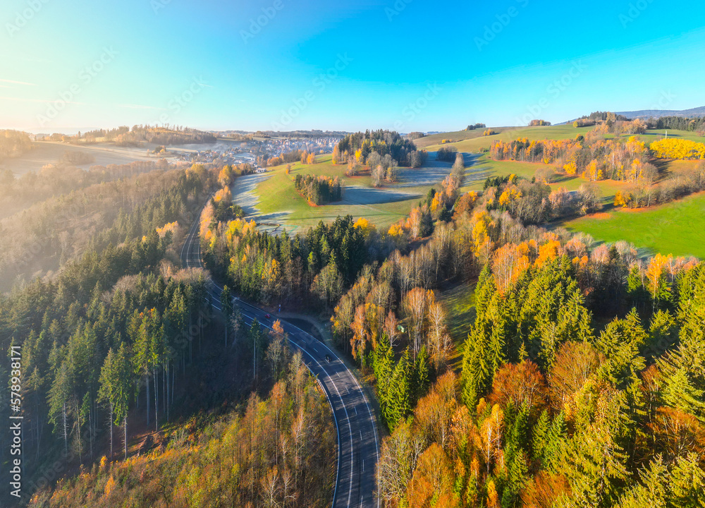 Aerial view of sunny, hilly and forested landscape illuminated by autumn sun. Asphalt road cutting through the scenery. Aerial view from drone.