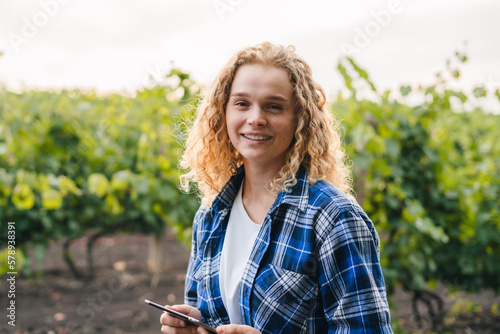 Close-up of happy young woman holding laptop standing in the vines hills examining grape crops and supply resources for human consumption. Checking plants and