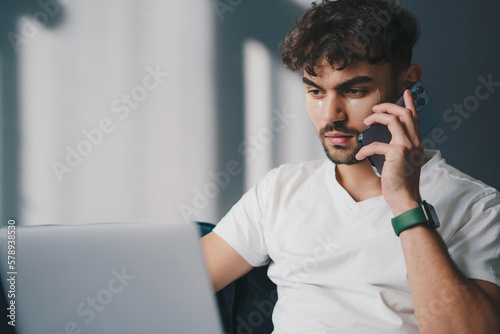 Young caucasian man relaxing at home on a sofa with his laptop computer having mobile phone conversation. Apartment interior. Internet network. People lifestyle