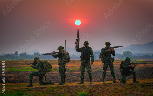 Fotografie Five soldiers are standing and aiming their weapons on the battlefield one of them is aiming their gun towards the sun during sunset in show to protect the land