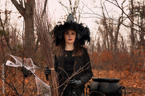 Beautiful woman in witches hat and costume holding broom near the cauldron in autumn forest. Halloween concept. Selective focus