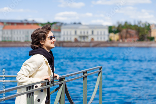 Happy beautiful young woman in white coat and sunglasses standing on embankment on sunny spring day gesticulates with happiness. The concept of Leisure and travel on vacation.