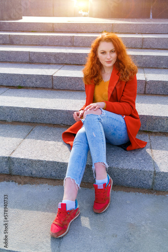 Young redhead woman in red coat yellow sweater and jeans sits on stairs in afternoon in sunny weather in the park in spring
