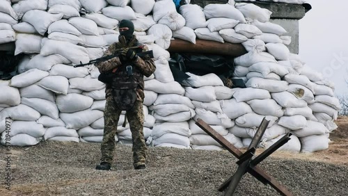 Soldier with weapon in military uniform stands in front of the barricades from sandbags and anti-tank hedgehog barriers. Military man on the roadblock. Combatant in full ammunition. Concept of war