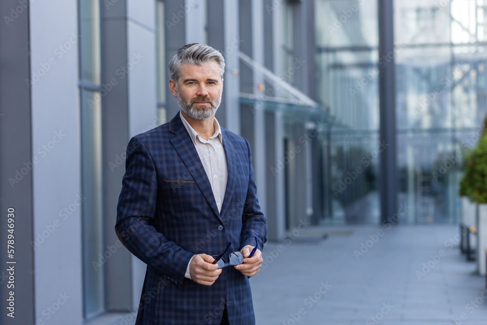 Portrait of successful mature man outside office building, businessman in business suit looking at camera thinking smiling while standing.