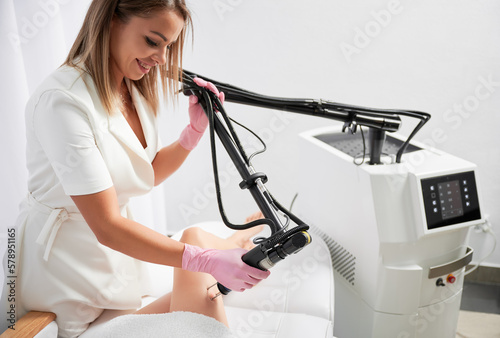 Doctor doing fractional treatment of skin on patient's leg by carbon dioxide laser in beauty clinic. Cosmetologist working with modern equipment in hardware cosmetology for laser skin rejuvenation. photo