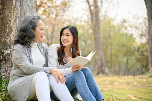 Happy Asian granddaughter reading a book with her grandmother under the tree in the park