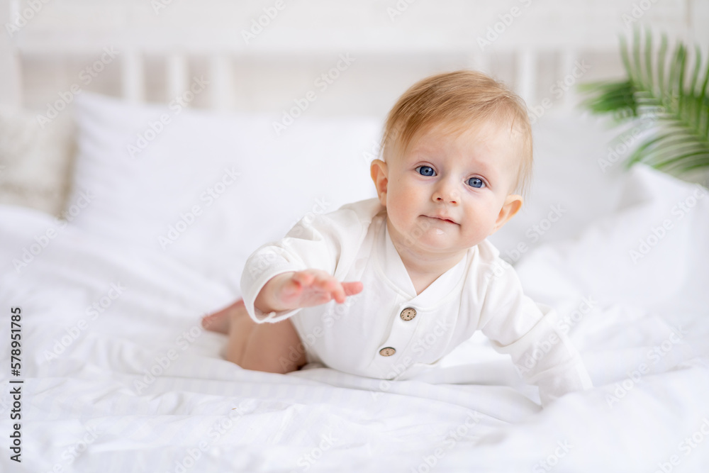 smiling baby 6 months old blond boy crawls on a white bed in a bright bedroom after sleeping in the morning in a cotton bodysuit, the concept of children's goods