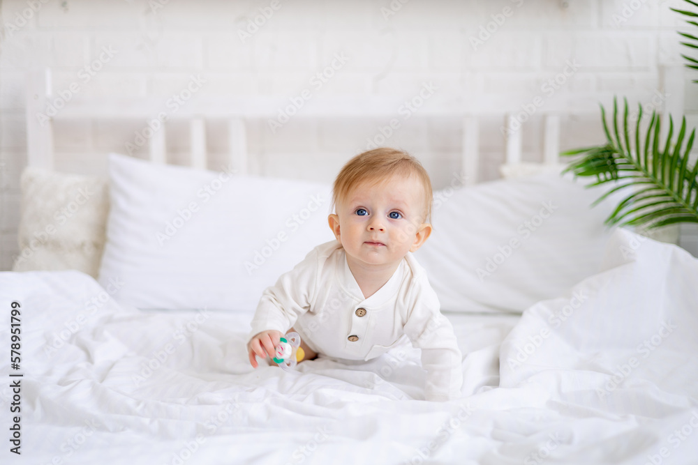 smiling baby 6 months old blond boy crawls on a white bed in a bright bedroom after sleeping in the morning in a cotton bodysuit, the concept of children's goods smiling baby 6 months old blond boy crawls on a white bed in a bright bedroom after sleeping in the morning in a cotton bodysuit, the concept of children's goods