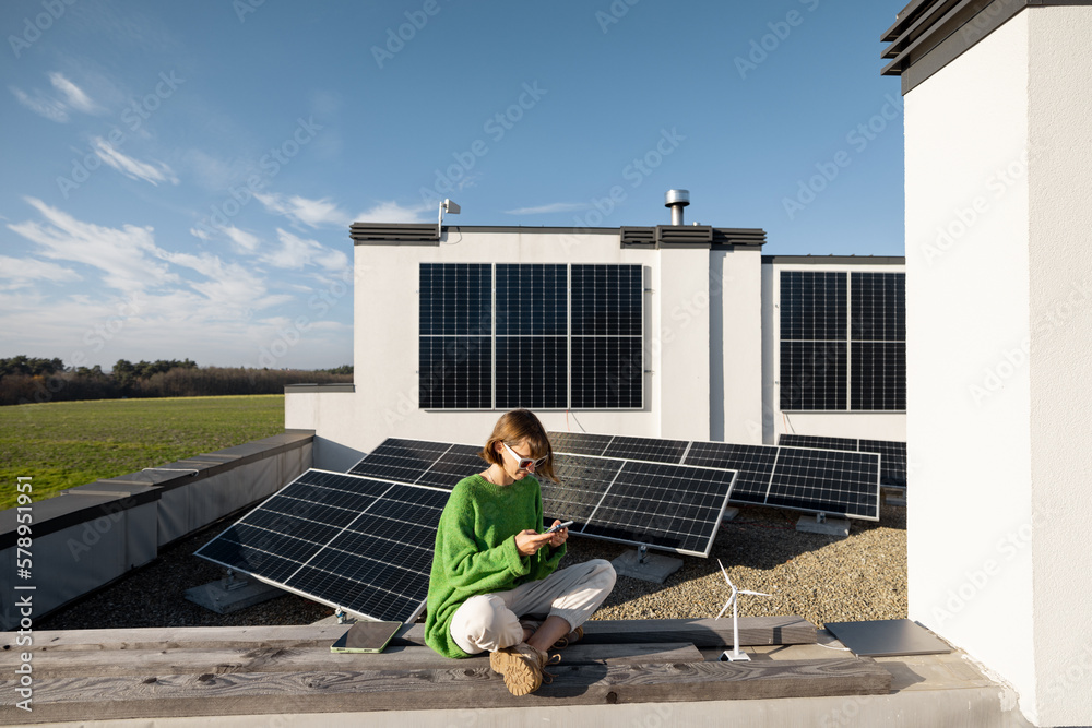Woman sits with phone on the rooftop of her house with a solar power plant installed on it. Happy owner of energy-independent household, sustainable lifestyle