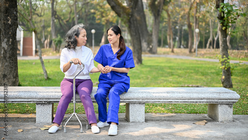 Asian female caregiver is talking while sitting on a bench in a park with an old disabled lady.