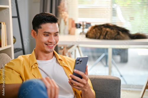 Handsome Asian man using his smartphone while relaxing on sofa in his living room.