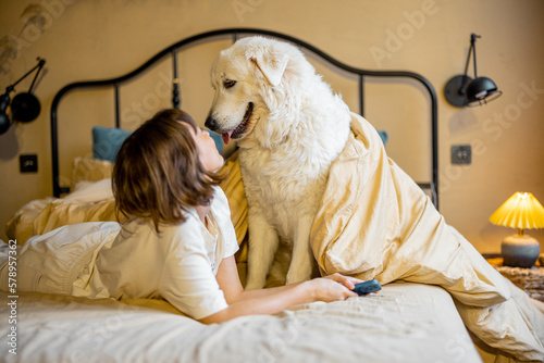 Young woman plays with her dog while lying together on bed at cozy bedroom in beige tones. Concept of friendship with pets and home coziness