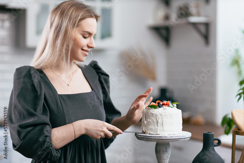 Pastry chef confectioner young caucasian woman in gray dress with knife cut slice cake on kitchen table. Cakes cupcakes and sweet dessert Scandinavian style