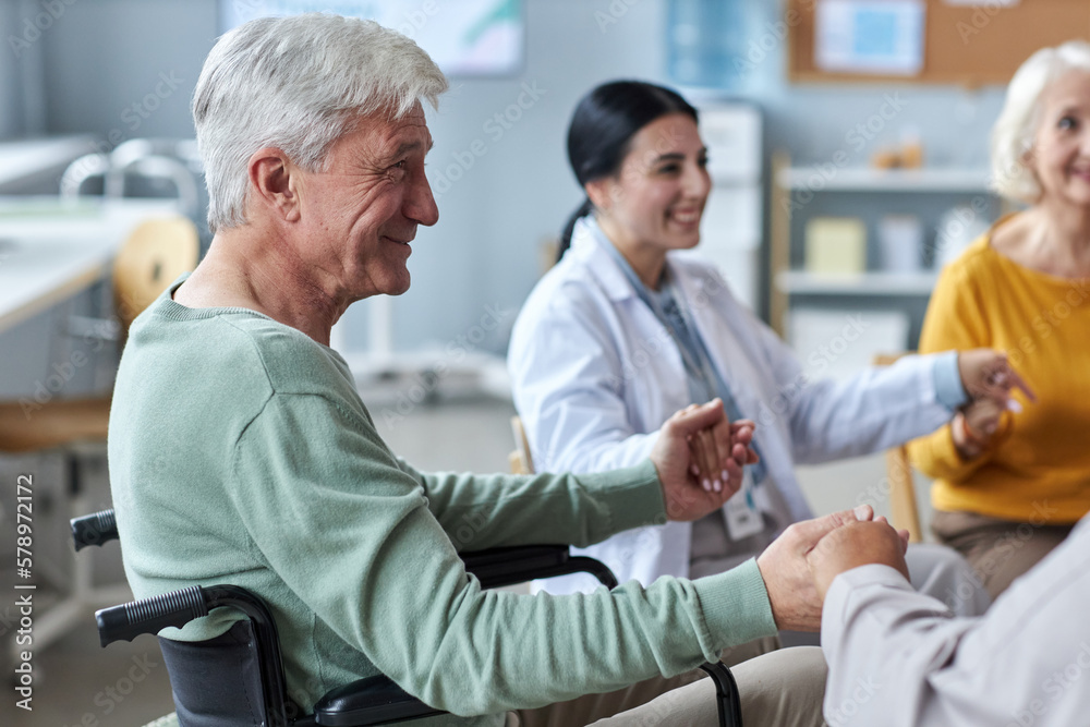 Side view portrait of smiling senior man holding hands with people in circle during support group session in retirement home Side view portrait of smiling senior man holding hands with people in circle during support group session in retirement home