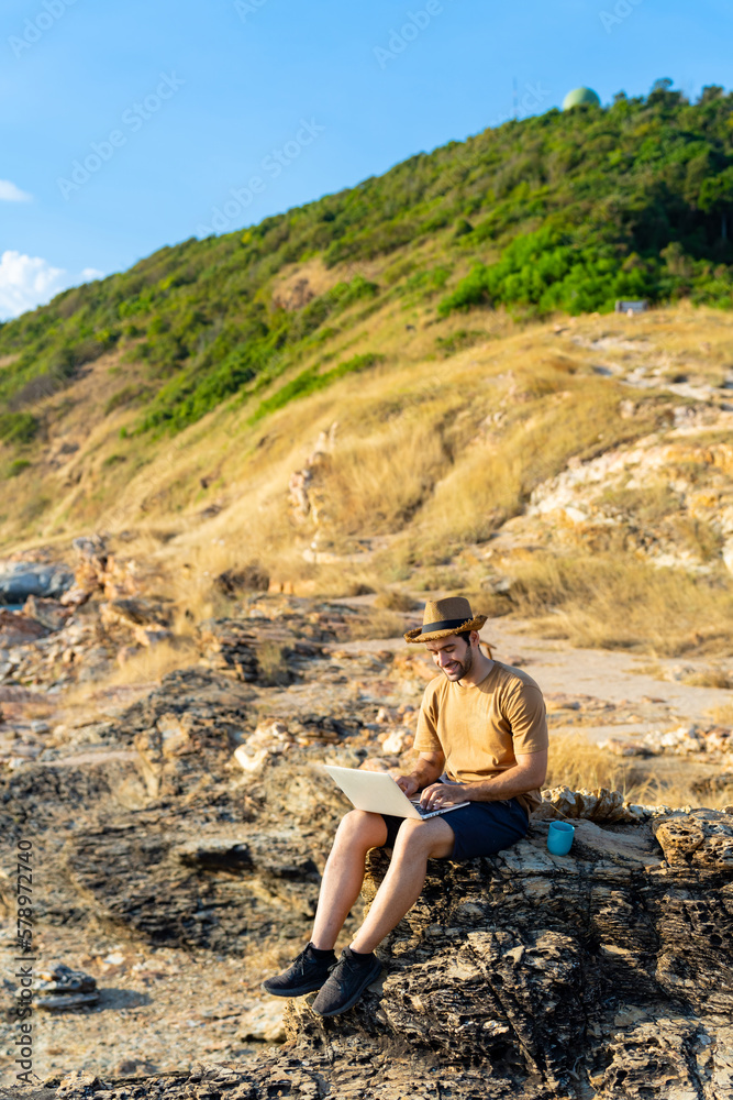 Caucasian man working outdoor on laptop computer and drinking coffee during sitting on coastline hill. Handsome guy hiking on mountain cliff on summer vacation. Digital nomad and solo travel concept.