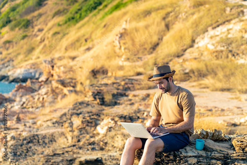 Caucasian man working outdoor on laptop computer and drinking coffee during sitting on coastline hill. Handsome guy hiking on mountain cliff on summer vacation. Digital nomad and solo travel concept. Caucasian man working outdoor on laptop computer and drinking coffee during sitting on coastline hill. Handsome guy hiking on mountain cliff on summer vacation. Digital nomad and solo travel concept.