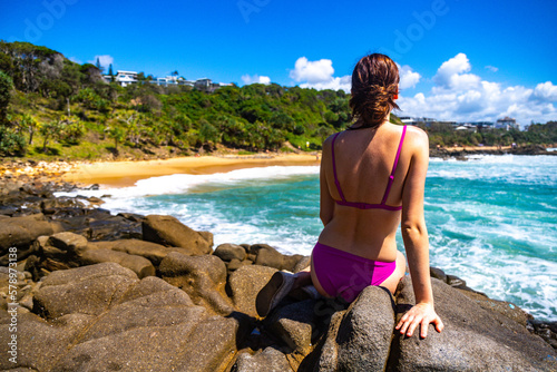 A beautiful girl in a pink bikini sits on a rock and enjoys the paradise scenery of small bays with golden sand and turquoise ocean in Sunshine Coast. Hidden gems in Queensland  Australia