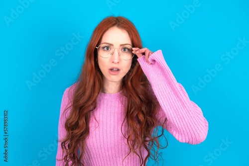 young woman wearing pink sweater over blue background having stunned and shocked look, with mouth open and jaw dropped exclaiming: Wow, I can't believe this. Surprise and shock