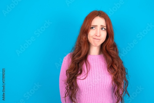 Amazed puzzled young woman wearing pink sweater over blue background , curves lips and has worried look, sees something awful in front. photo