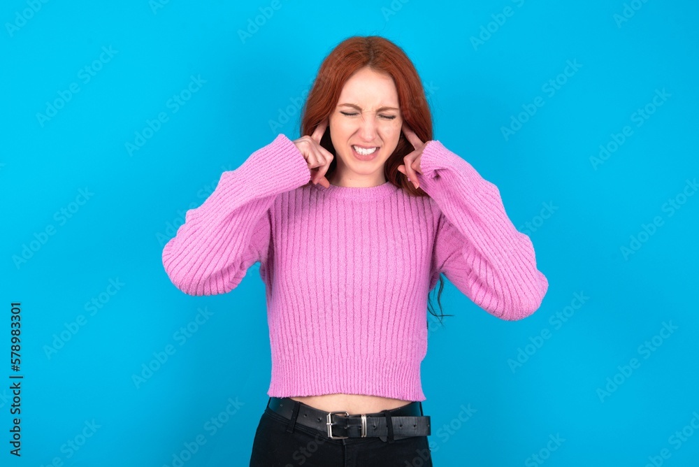 Stop making this annoying sound! Unhappy stressed out young woman wearing pink sweater over blue background making worry face, plugging ears with fingers, irritated with loud noise. Stop making this annoying sound! Unhappy stressed out young woman wearing pink sweater over blue background making worry face, plugging ears with fingers, irritated with loud noise.