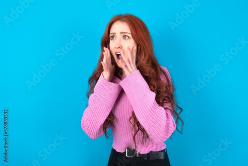 young woman wearing pink sweater over blue background with shocked facial expression, holding hands near face, screaming and looking sideways at something amazing. © Jihan