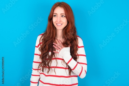 Joyful young woman wearing striped sweater over blue background expresses positive emotions recalls something funny keeps hand on chest and giggles happily. photo