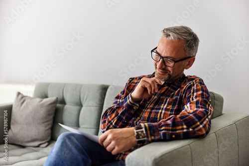 A focused businessman looking at the notes he wrote on paper, sitting on the couch.