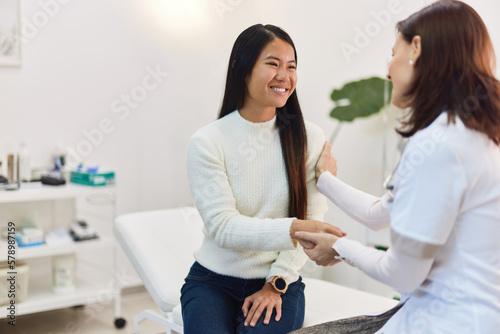 A smiling Asian woman patient sitting with a female doctor in the examination room.