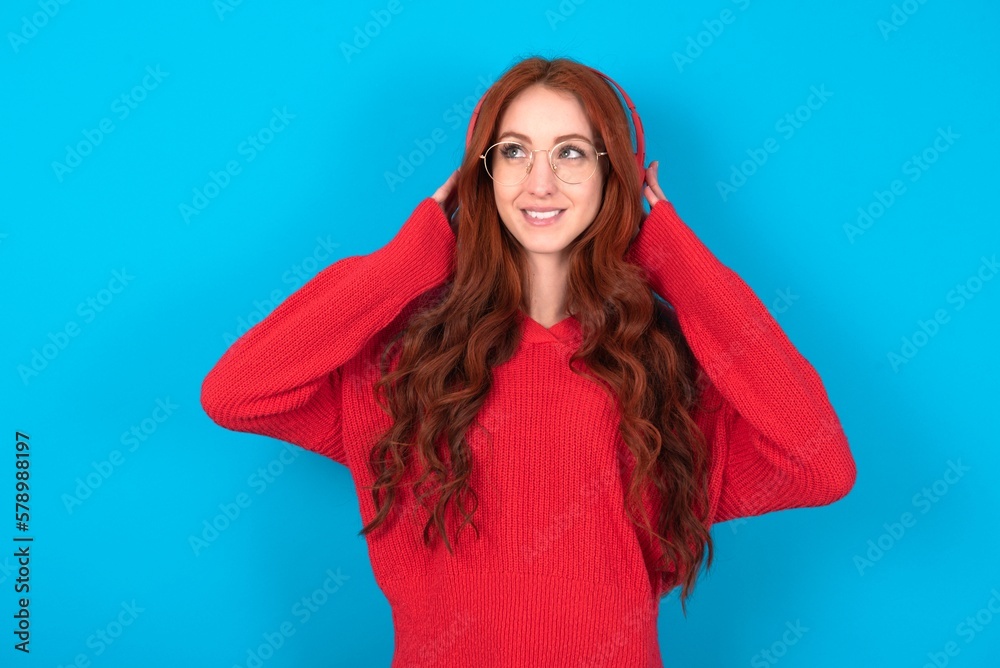 Joyful young woman wearing red sweater over blue background sings song keeps hand near mouth as if microphone listens favorite playlist via headphones Joyful young woman wearing red sweater over blue background sings song keeps hand near mouth as if microphone listens favorite playlist via headphones
