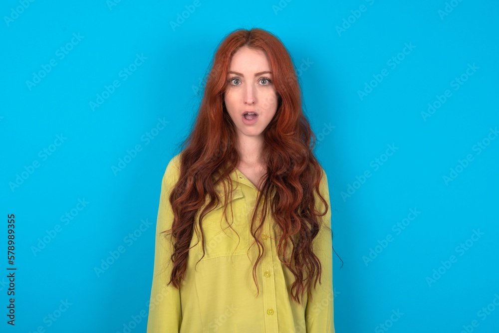 Fototapeta premium Oh my God. Surprised young woman wearing green sweater over blue background stares at camera with shocked expression exclaims with unexpectedness,