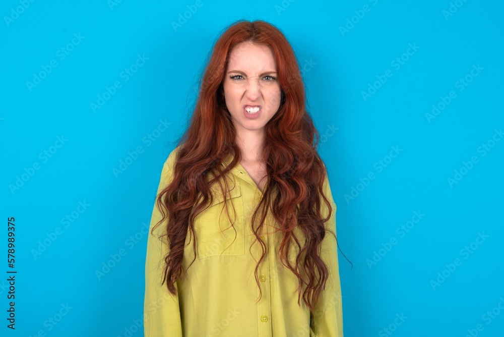 young woman wearing green sweater over blue background keeps teeth clenched, frowns face in dissatisfaction, irritated because of much duties. young woman wearing green sweater over blue background keeps teeth clenched, frowns face in dissatisfaction, irritated because of much duties.