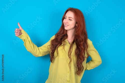 young woman wearing green sweater over blue background Looking proud, smiling doing thumbs up gesture to the side. Good job!