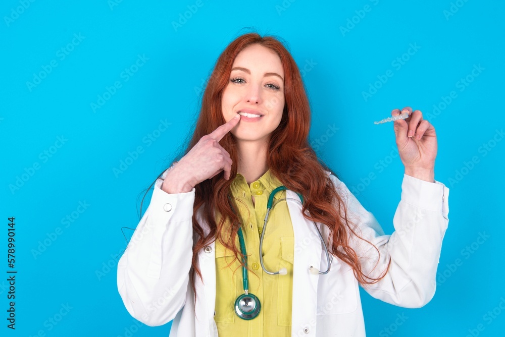 young doctor woman wearing medical uniform over blue background holding an invisible aligner and pointing to her perfect straight teeth. Dental healthcare and confidence concept. young doctor woman wearing medical uniform over blue background holding an invisible aligner and pointing to her perfect straight teeth. Dental healthcare and confidence concept.