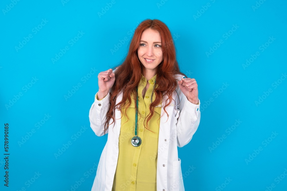 young doctor woman wearing medical uniform over blue background clenches fists and awaits for something nice happened looks away bites lips and waits announcement of results young doctor woman wearing medical uniform over blue background clenches fists and awaits for something nice happened looks away bites lips and waits announcement of results