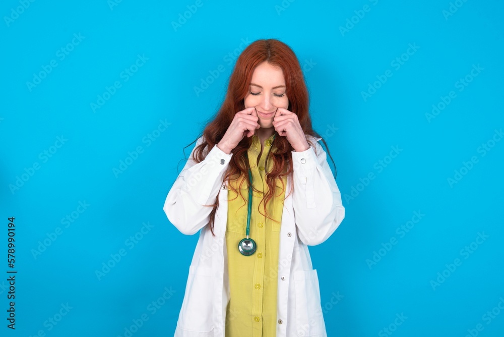 Pleased young doctor woman wearing medical uniform over blue background with closed eyes keeps hands near cheeks and smiles tenderly imagines something very pleasant Pleased young doctor woman wearing medical uniform over blue background with closed eyes keeps hands near cheeks and smiles tenderly imagines something very pleasant