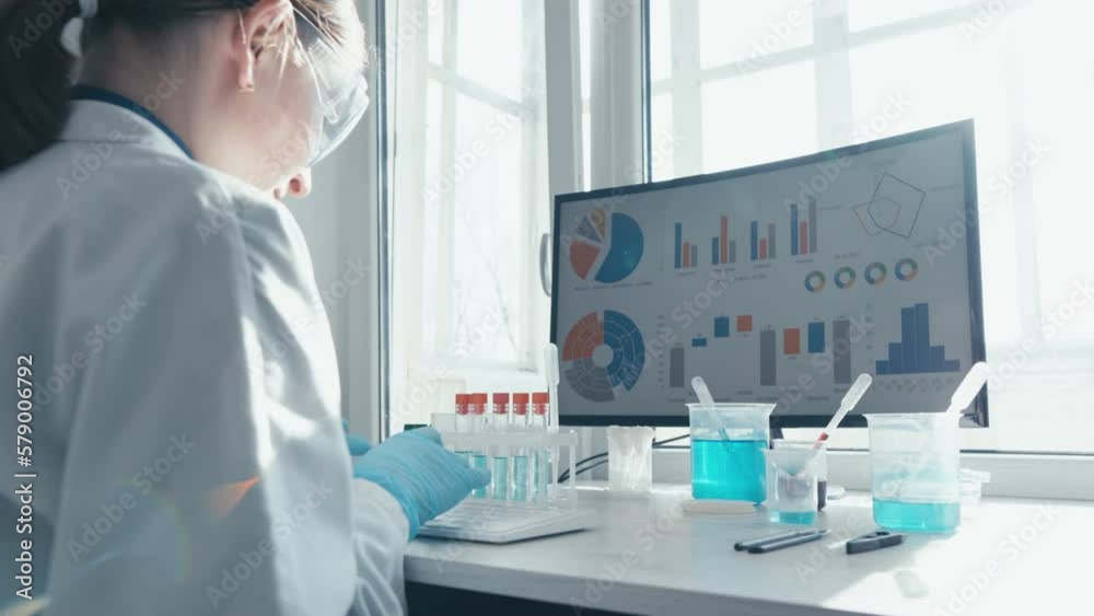 A female researcher sits at a workplace in a laboratory, behind a personal computer monitor. Against the background research statistics. Pharmaceutical medical worker in protection works at keyboard. A female researcher sits at a workplace in a laboratory, behind a personal computer monitor. Against the background research statistics. Pharmaceutical medical worker in protection works at keyboard.