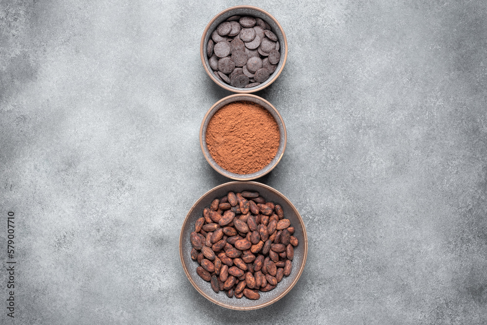 Cocoa beans, cocoa powder and chocolate in bowls on a gray background. Top view, vertical border. Cocoa beans, cocoa powder and chocolate in bowls on a gray background. Top view, vertical border.