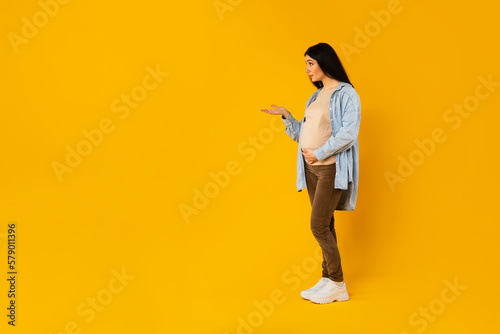 Pregnant woman looking at free space, holding open palm while standing isolated over yellow background, full length