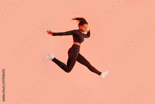Studio shot of fit young black woman jumping on peach neon light studio background, free space