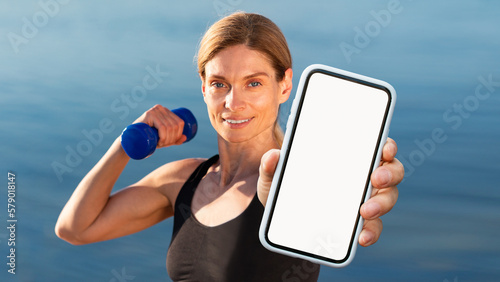 Sporty Middle Aged Woman Training With Dumbbells Outdoors And Showing Blank Smartphone photo