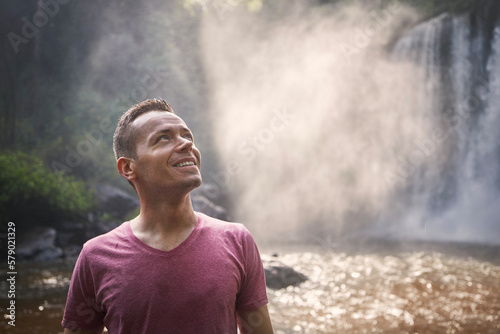 Portrait of traveler against high waterfall. Happy man in tropical nature in Cambodia..