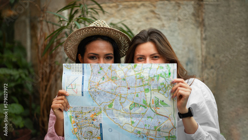 Smiling young african american and european women in hat looking for route peeking out from behind map photo