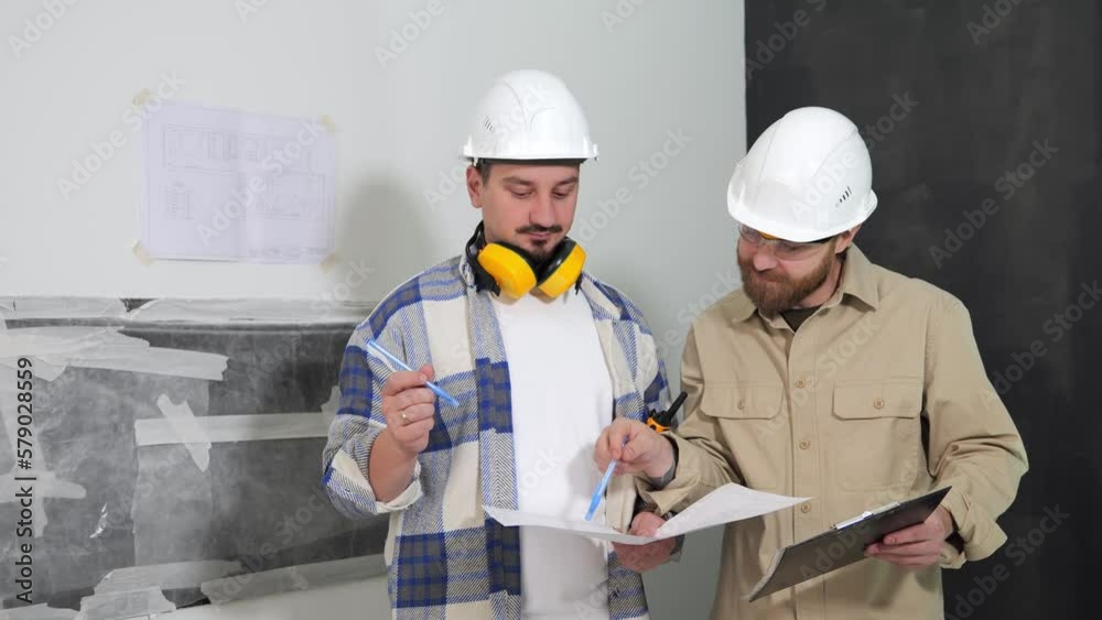 Two engineers in white helmets at a construction site are holding a drawing in their hands and discussing it. Point the handles to the drawing and to the objects in the room. High quality 4k footage Two engineers in white helmets at a construction site are holding a drawing in their hands and discussing it. Point the handles to the drawing and to the objects in the room. High quality 4k footage