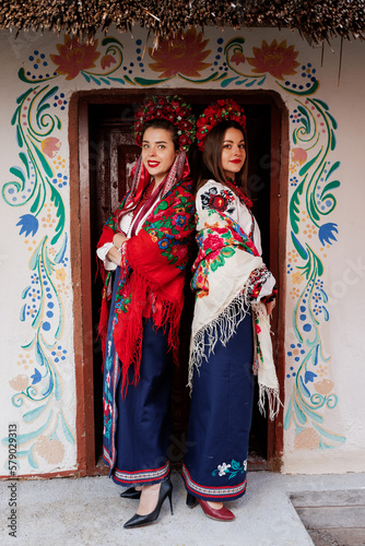 Two Ukrainian women in traditional ethnic clothing and floral red wreath on background of decorated hut. Ukrainian national embroidered dress call vyshyvanka. Pray for Ukraine