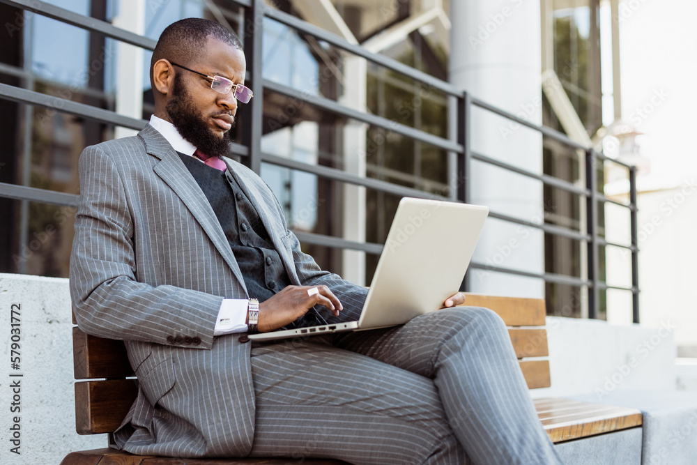 A dark-skinned man in a business suit is using a laptop outdoors. Digital education and remote work A dark-skinned man in a business suit is using a laptop outdoors. Digital education and remote work